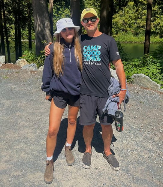 Thomas Mould is standing with his arm around his daughter in a wooded area at Camp Goodtimes