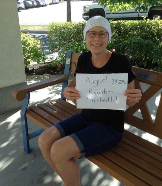 Vickie Lachetta assise sur un banc public, brandissant une pancarte sur laquelle est écrit en anglais « 24 août 2016 : Finie la radiothérapie!!! »