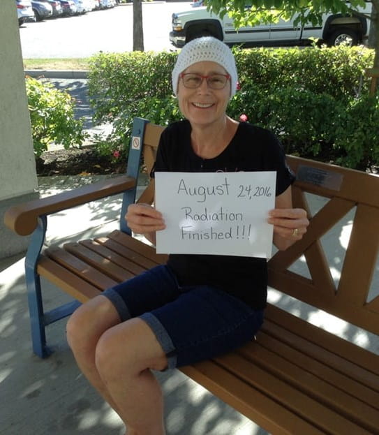 Vickie Iachetta, sitting on a park bench, holding up a sign that reads “August 24, 2016 Radiation Finished!!”