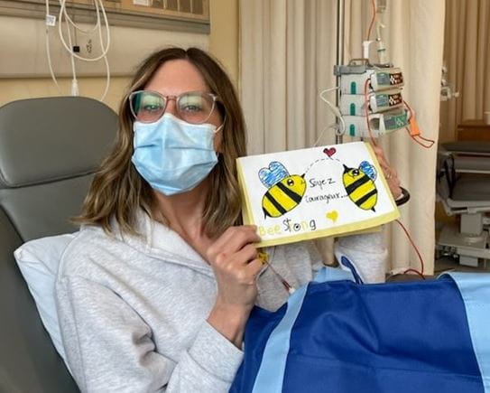 Amélie sitting in a hospital bed and is showing a drawing with the words “be strong”