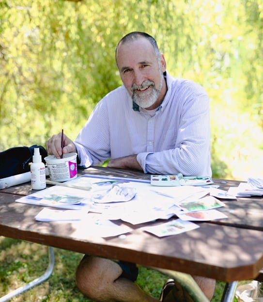 Angus Pratt assis à une table sous les arbres, les bras croisés.