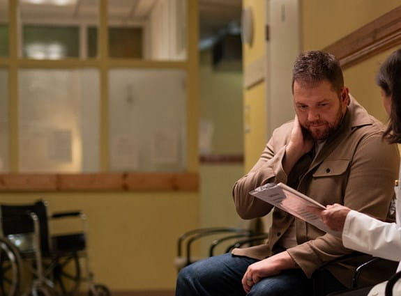 Un médecine tient un dossier médical et parle à une personne assise dans un couloir d’hôpital.