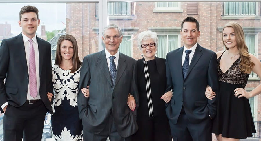 A group of people in formal clothing smiling and posing with their arms linked in front of a window.