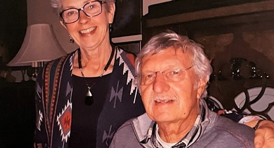 A husband and wife smiling. Both are in casual attire and wear glasses.