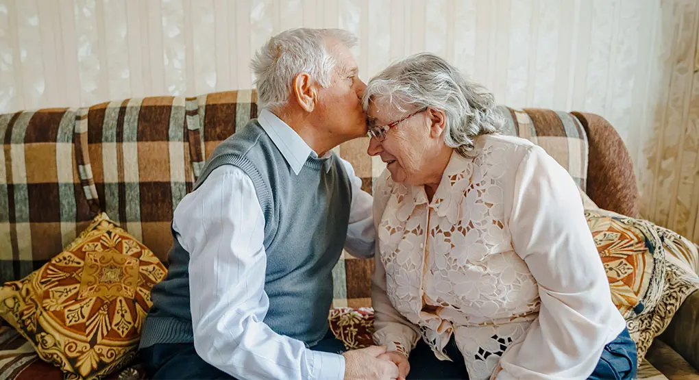 An elderly couple sits on a sofa, leaning towards each other and holding hands while the man is kissing his wife on the forehead.