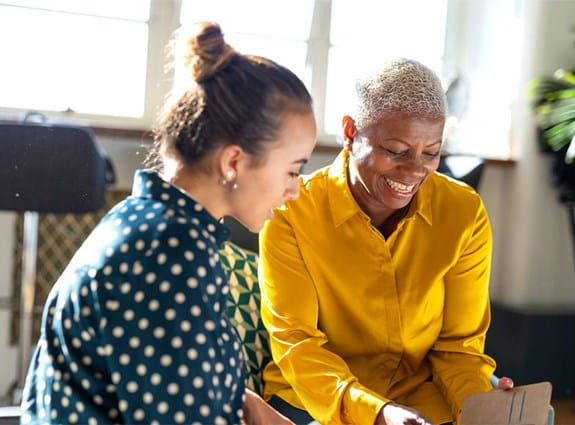 Une femme assise regarde son téléphone mobile en souriant. Une autre femme assise à côté d’elle regarde le téléphone elle aussi.