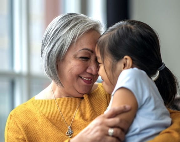 Une femme tient sa petite-fille dans ses bras. Elles pressent leurs fronts l’un contre l’autre en se souriant.