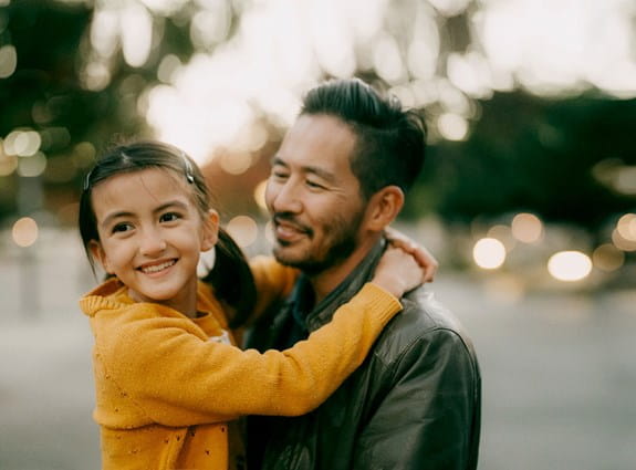 Un homme debout sourit en regardant sa fille. Elle a les bras enroulés autour de ses épaules et il la tient dans ses bras. Elle sourit en regardant au loin.