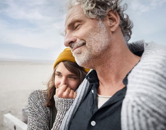 Un homme se tient debout, les yeux fermés, sur la plage. Une femme se tient à ses côtés, appuyée sur son épaule, la main sur la bouche.